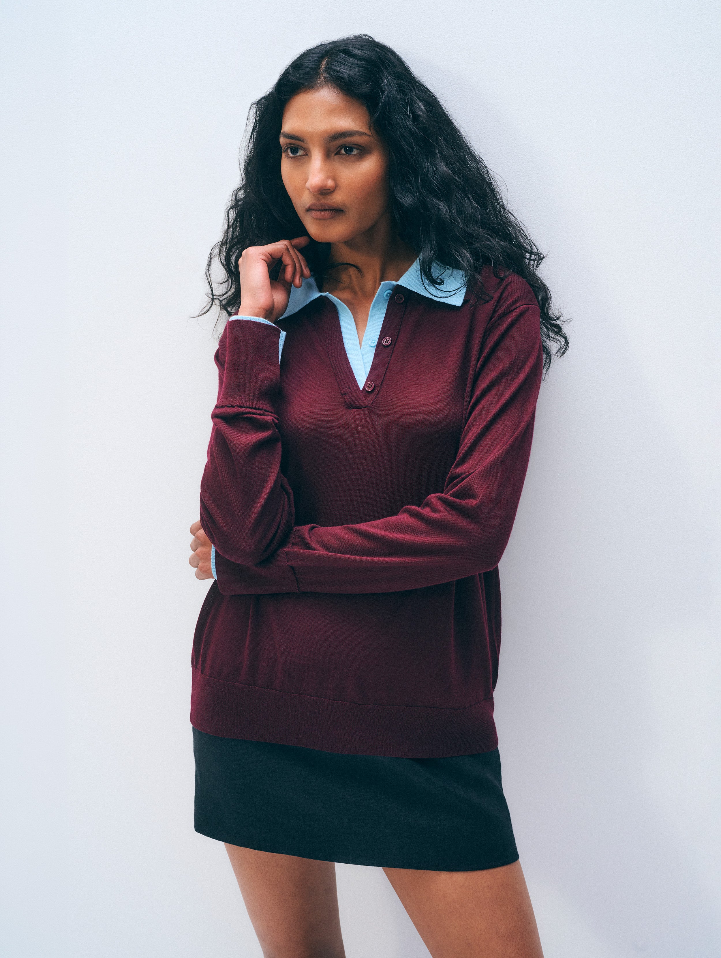 A woman modeling a long-sleeve polo shirt in maroon with a light blue collar and cuffs, paired with a black skirt, against a plain light gray background.