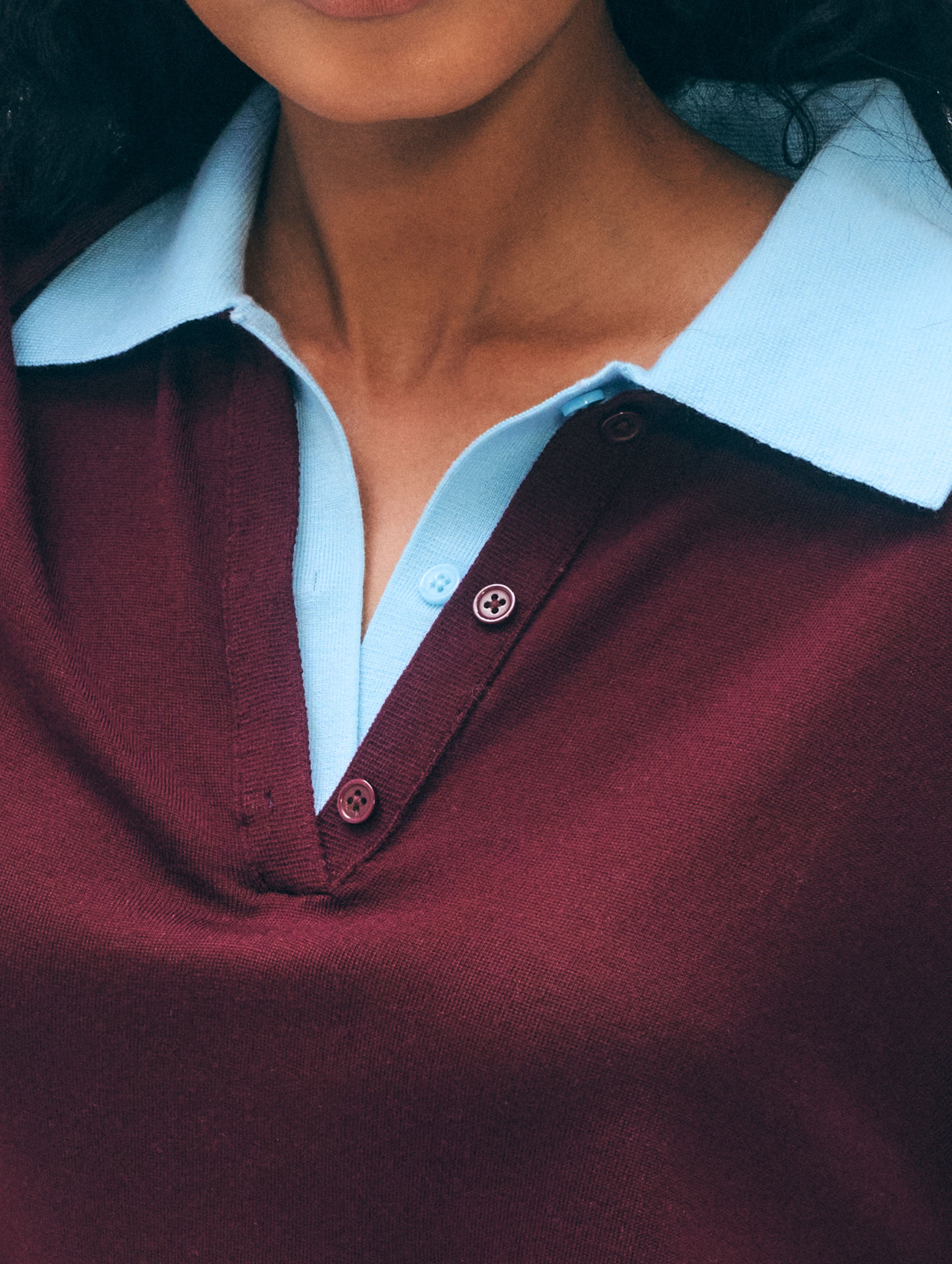 Close-up of a person wearing a burgundy knit polo shirt layered over a light blue collared shirt. The photo is tightly cropped from the lower face to the chest, showing the collar, buttons, and knit texture.