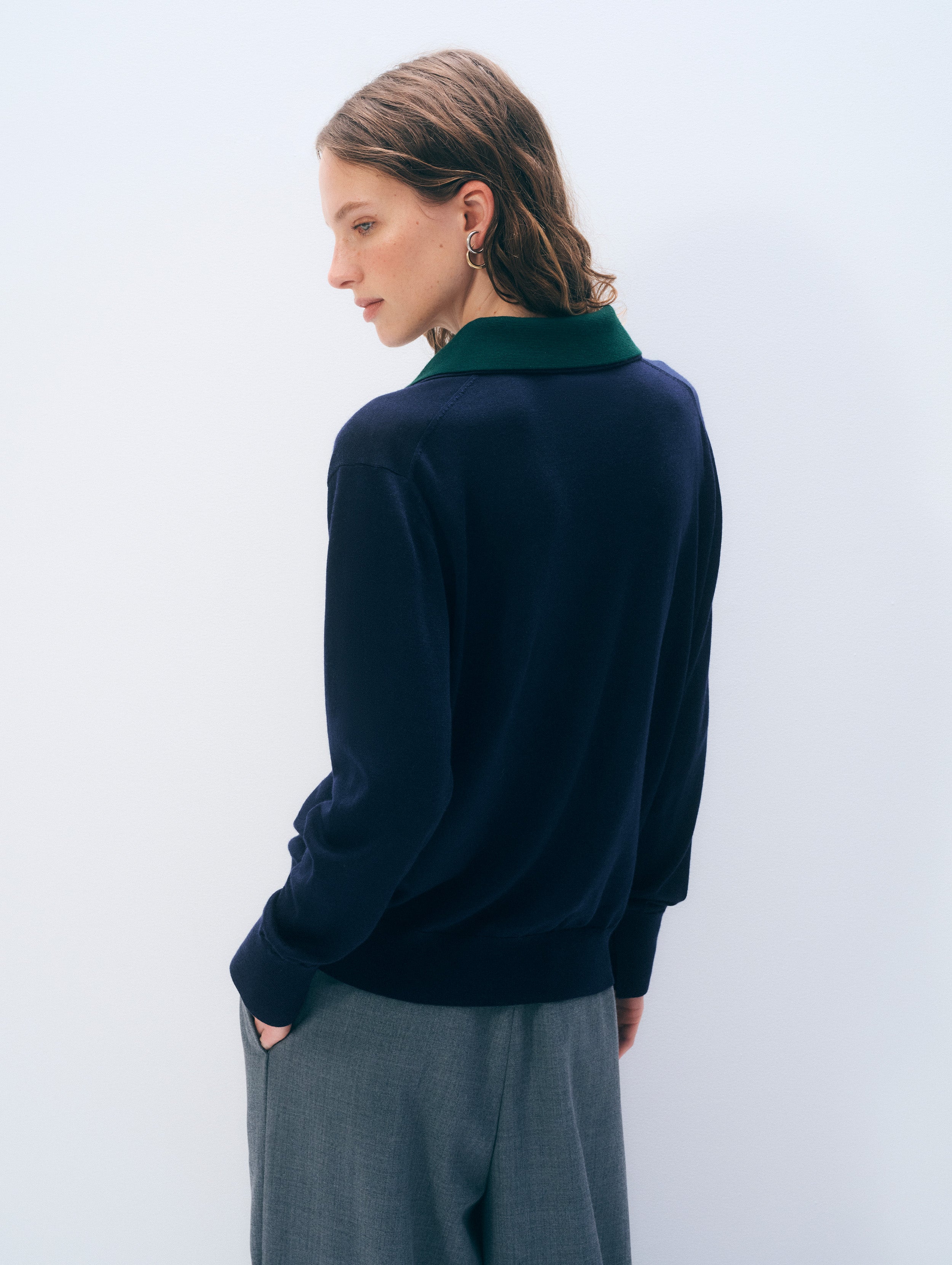 A woman shown from the back wearing a navy blue knit sweater with a contrasting dark green collar, paired with gray trousers, against a plain light gray background.