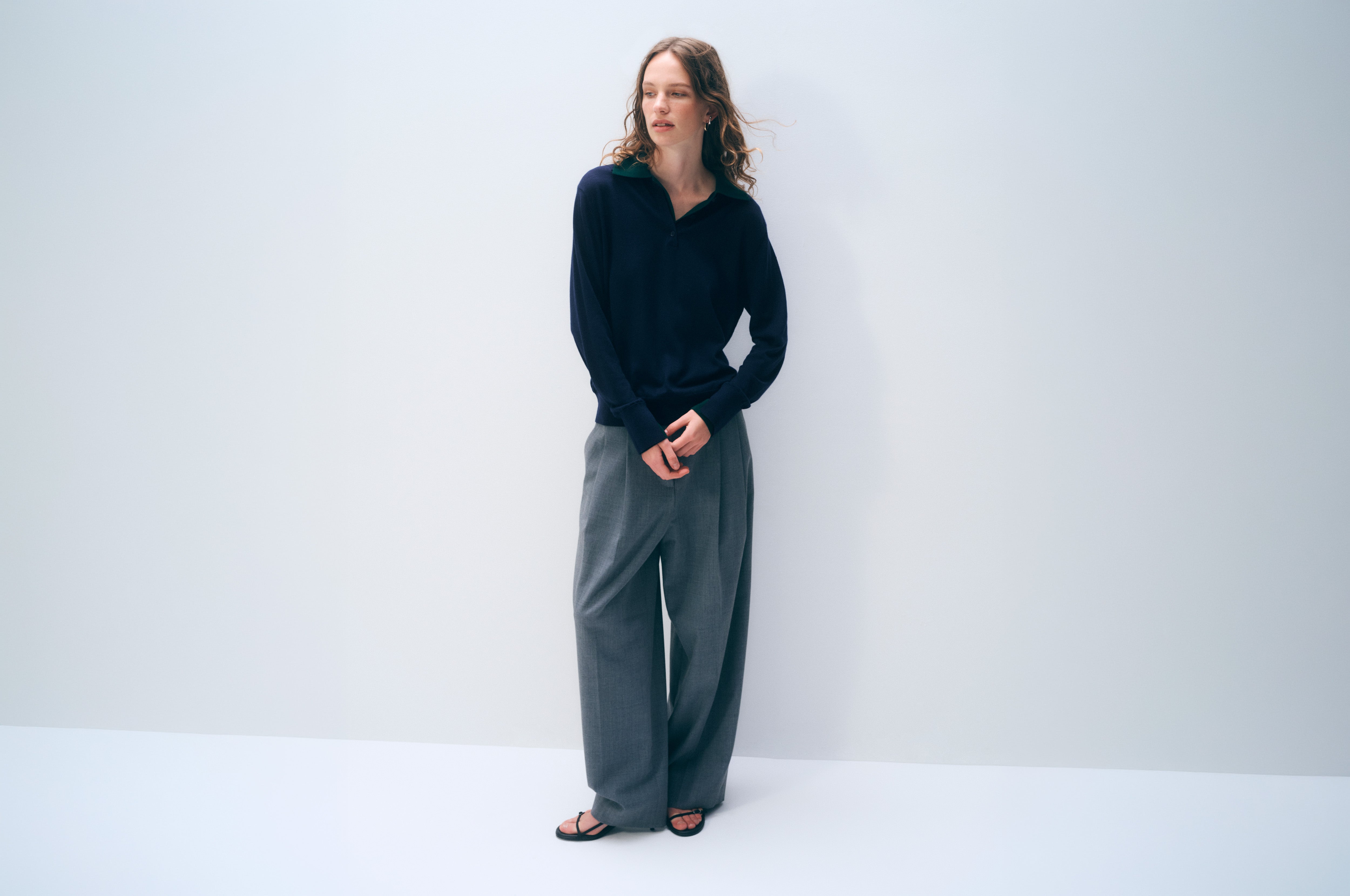 A woman stands against a plain light gray studio wall wearing a dark navy long-sleeve knit top, loose gray wide-leg pants, and black sandals.