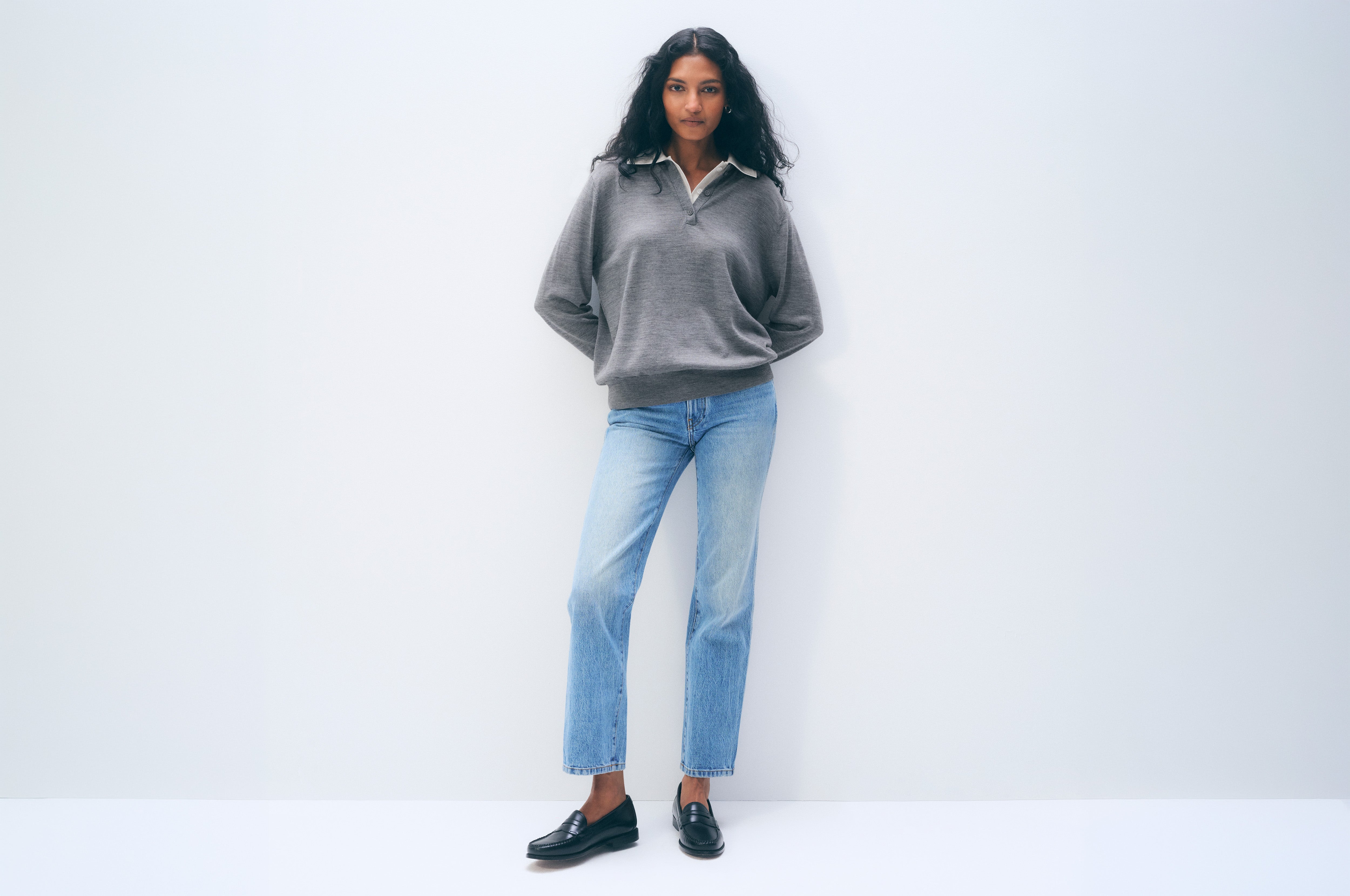 A woman stands against a plain white studio wall wearing a gray long-sleeve polo-style sweater, light blue straight-leg jeans, and black loafers.