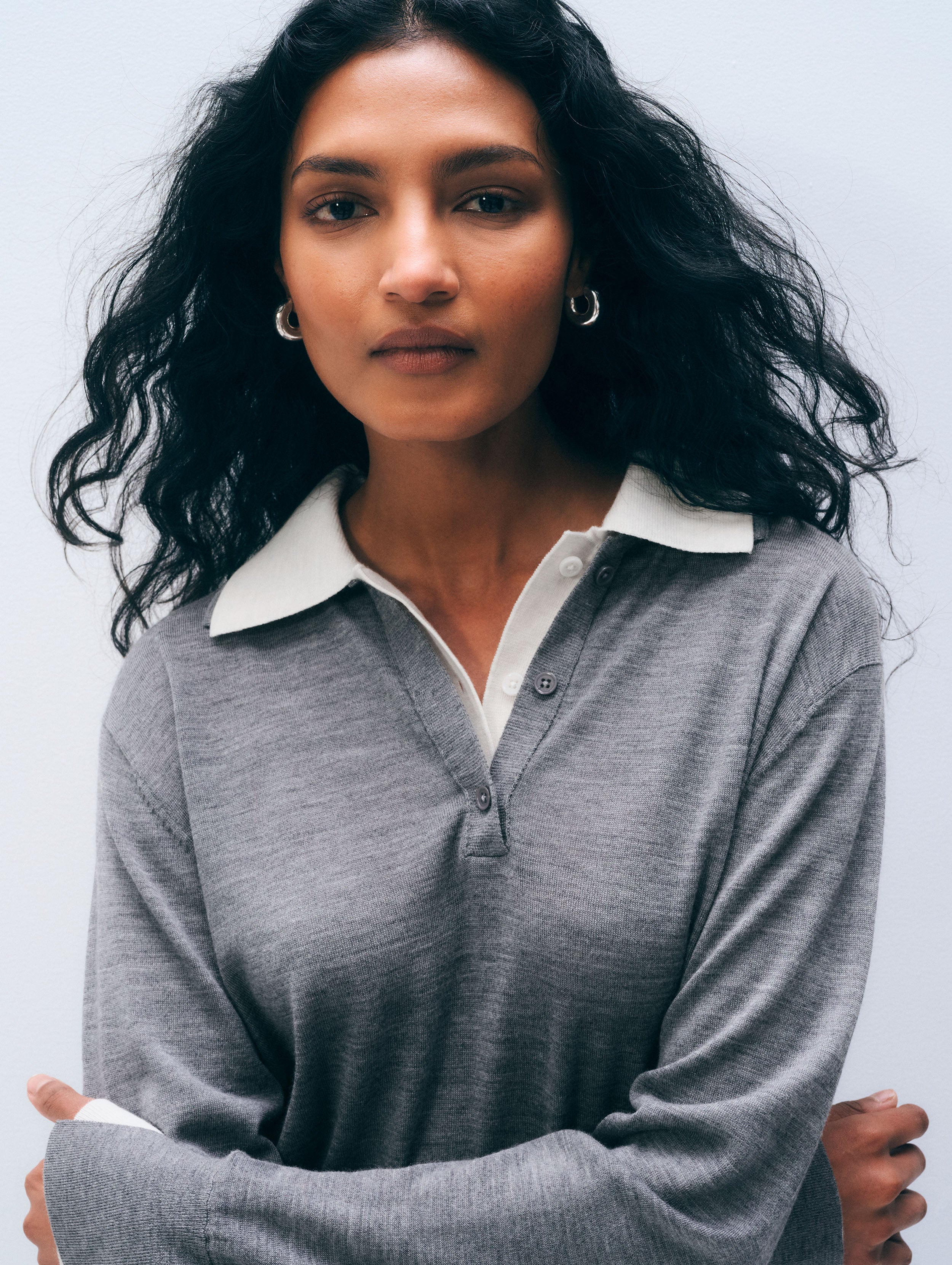 A woman with long, wavy black hair wearing a heather gray long-sleeve polo shirt with a contrasting white collar, posed against a light gray studio background.
