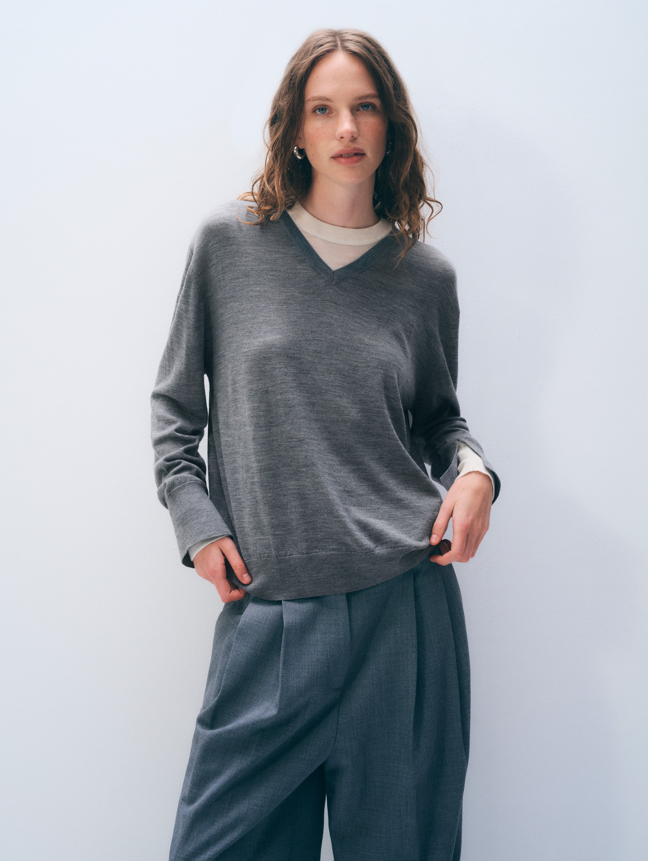 A woman modeling a dark gray long-sleeve V-neck knit sweater over a cream undershirt, paired with dark gray wide-leg trousers, against a light gray studio background.