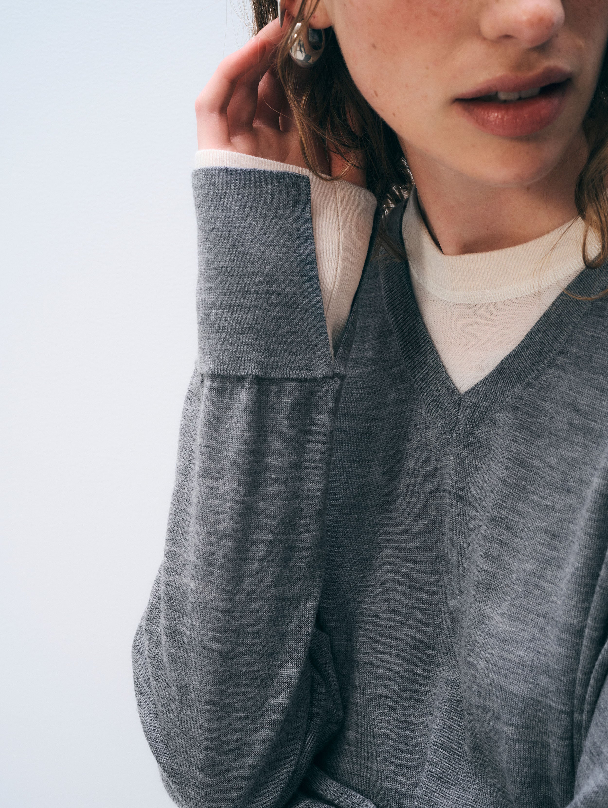 Close-up of a person wearing a heather gray knit sweater over a cream V-neck undershirt, with part of the face and hand visible against a pale blue background.