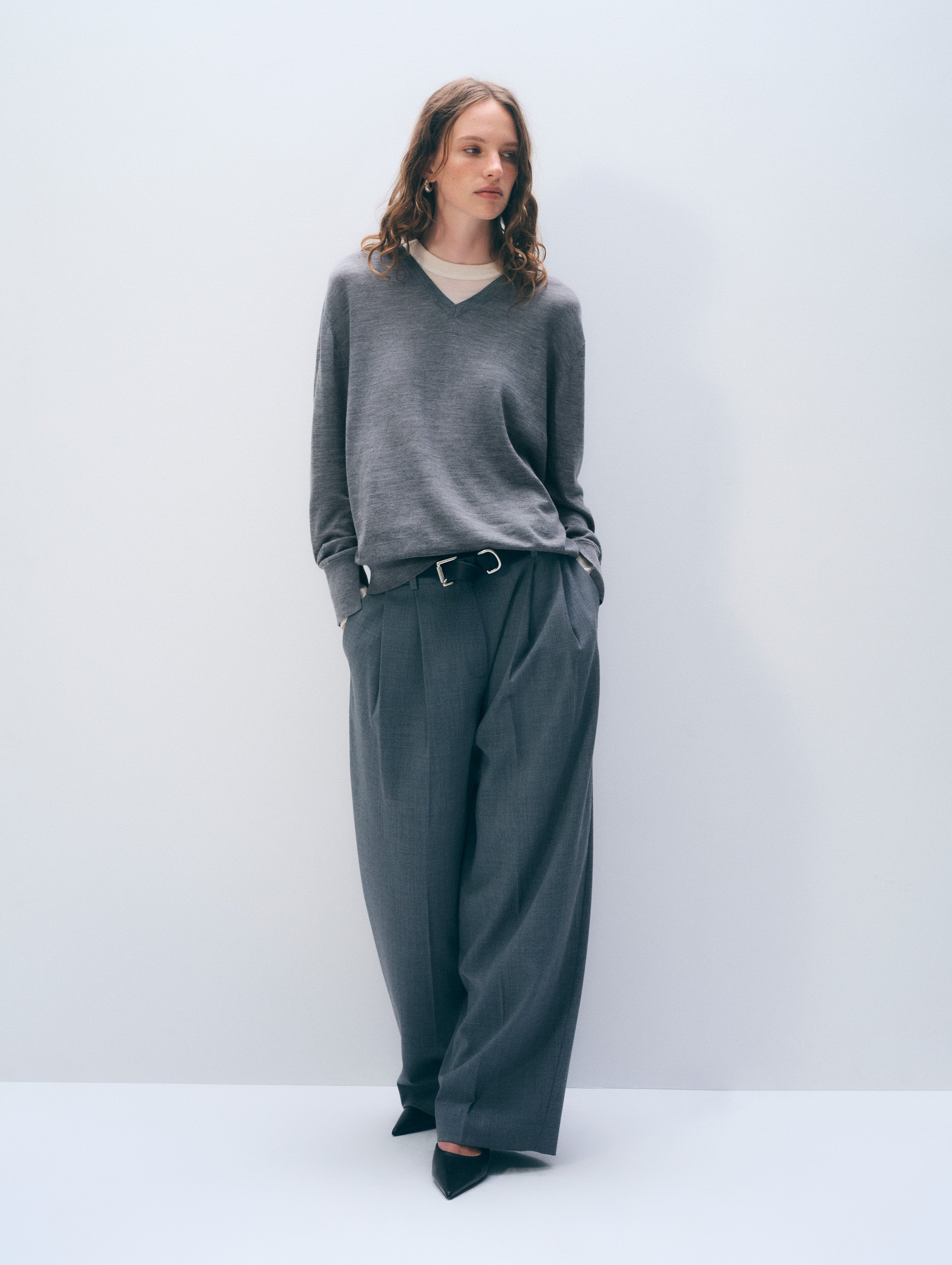 A woman stands against a light gray studio background wearing a gray V-neck sweater, dark gray wide-leg trousers with a belt, and black pointed shoes.