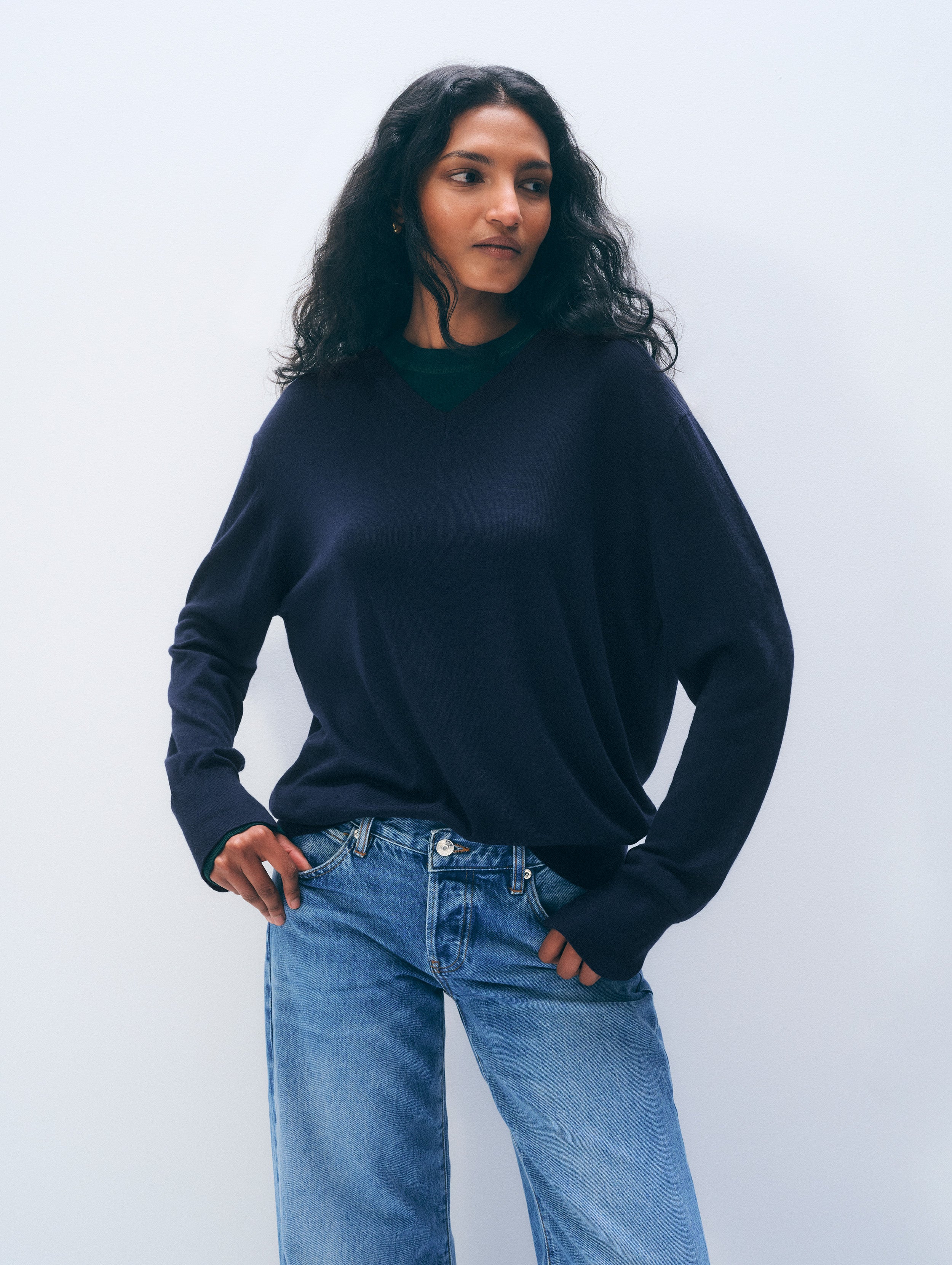 A woman wearing a dark navy blue knit sweater and light blue jeans, standing against a light gray studio background.