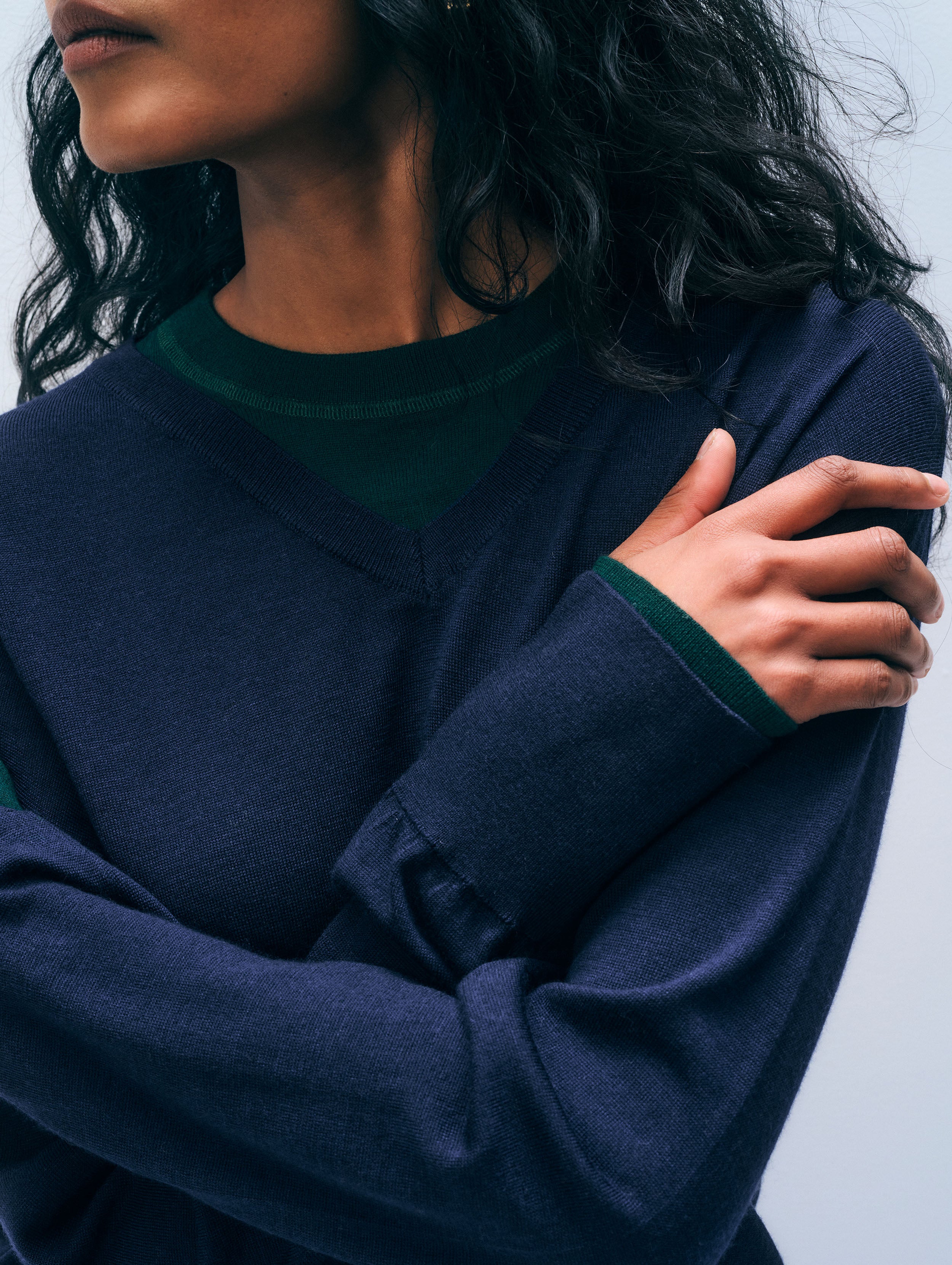A close-up of a person wearing a navy blue long-sleeve V-neck sweater with dark green ribbed trim at the collar and cuffs, with one arm crossed over the other.