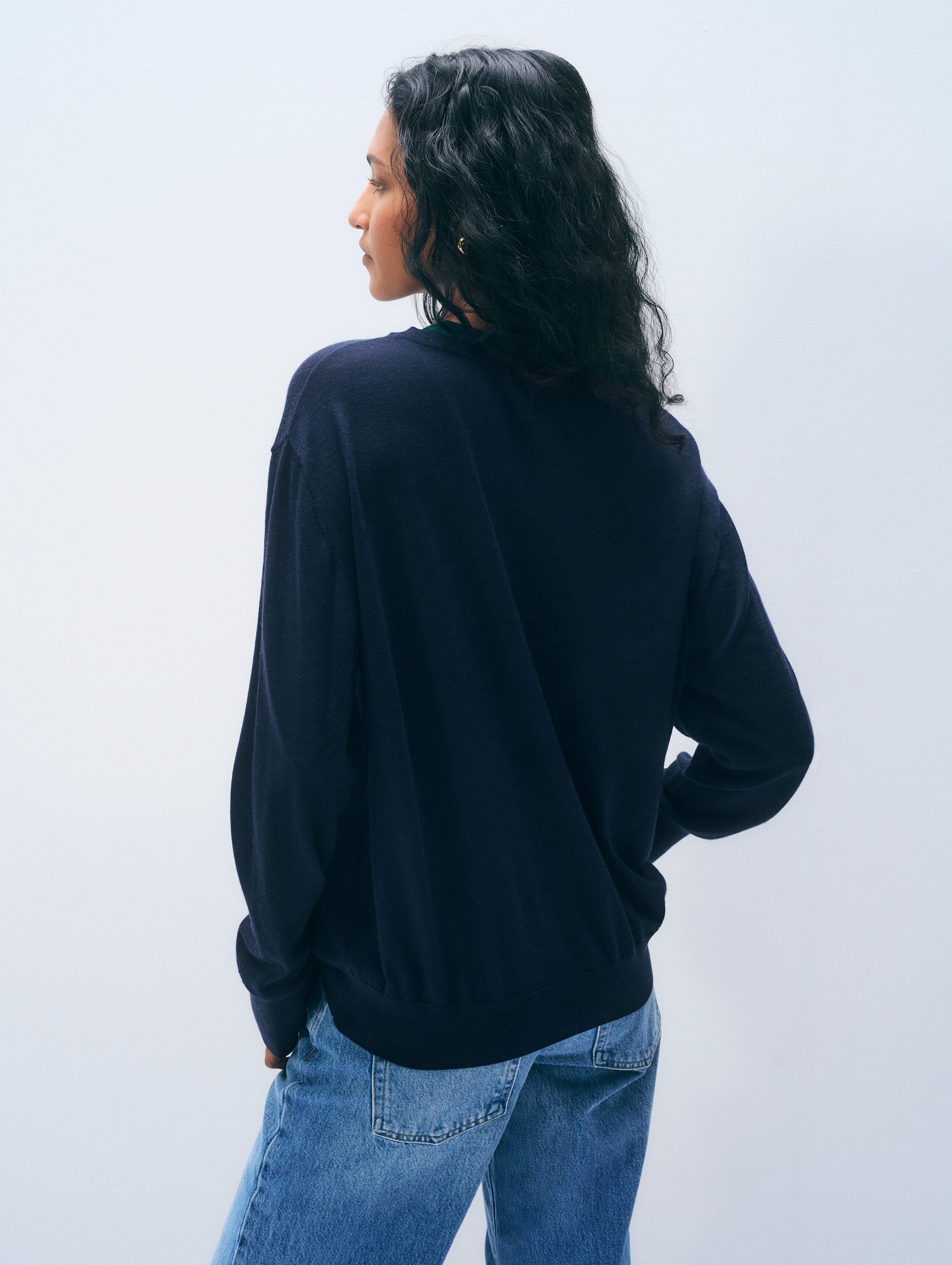A woman with long dark curly hair is shown from the back wearing a loose-fitting black long-sleeve sweater and blue jeans against a plain light gray studio background.