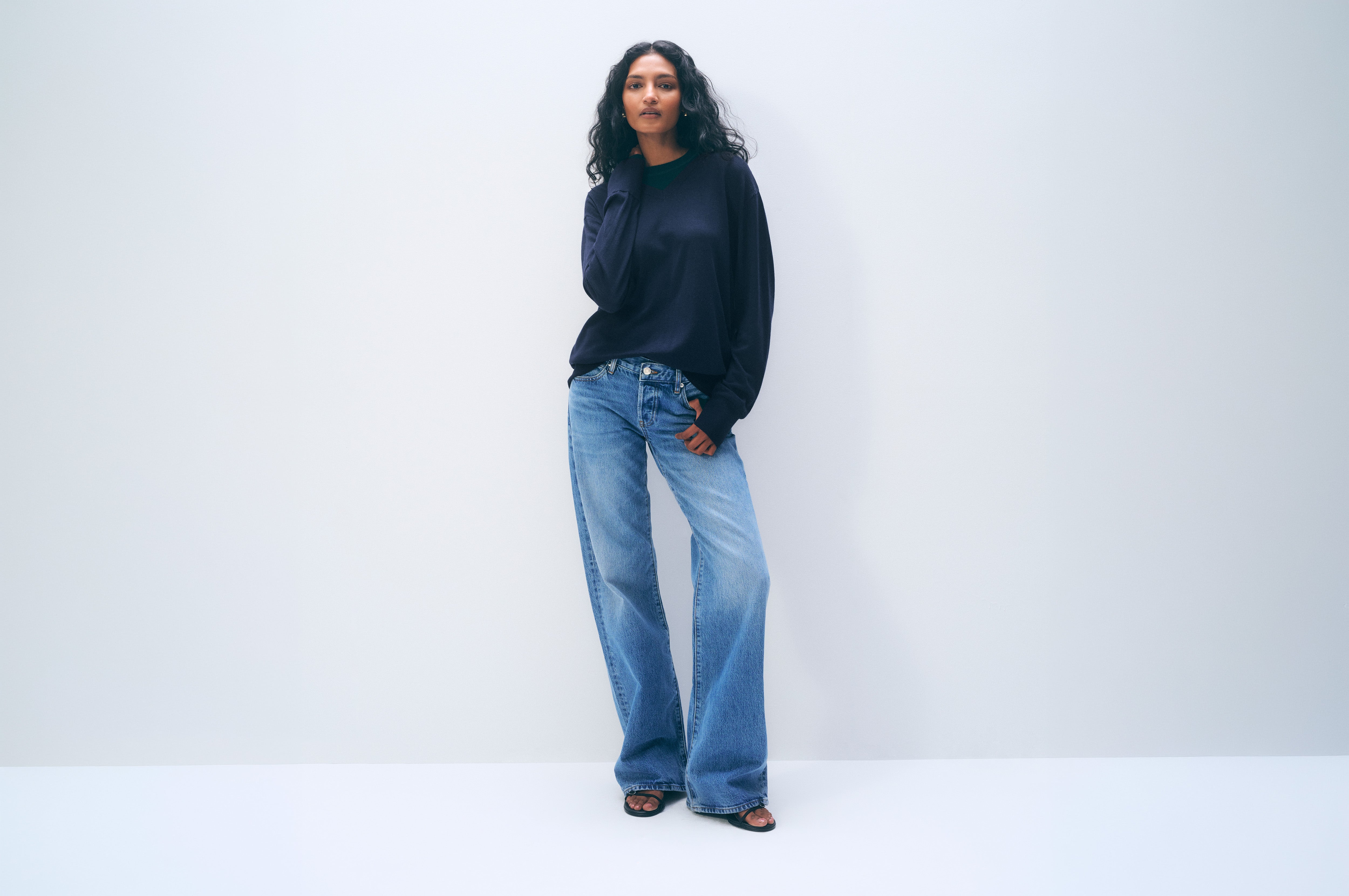 A woman stands against a plain light gray studio wall wearing a dark navy long-sleeve top, wide-leg light blue jeans, and dark sandals.