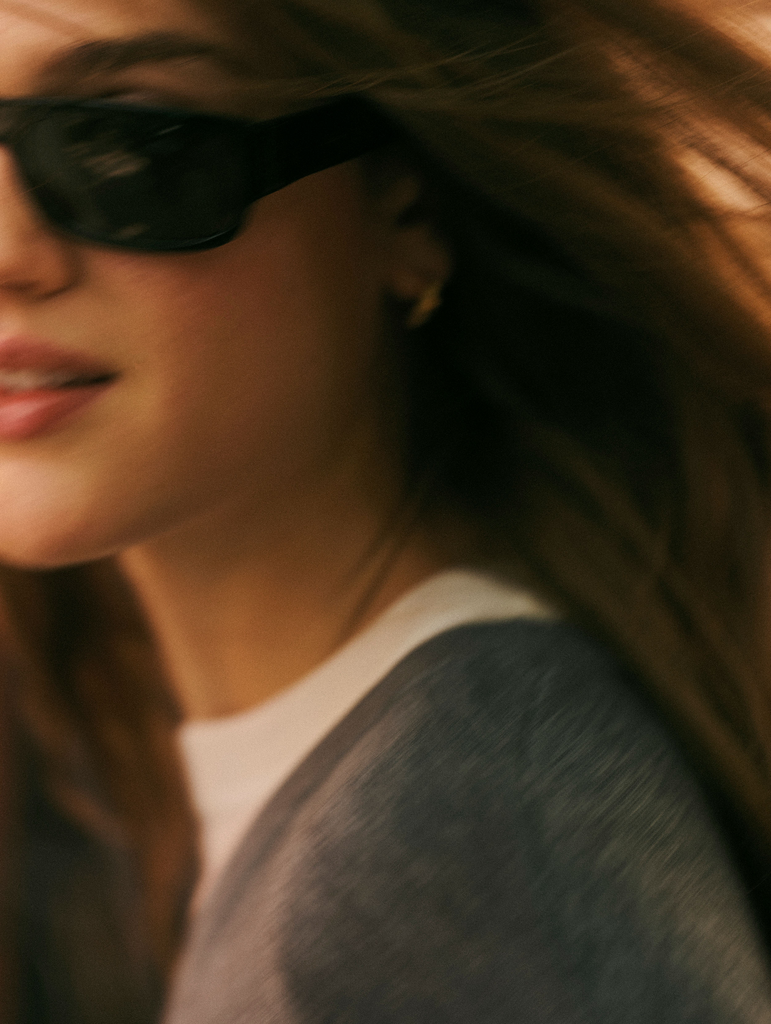 A close-up lifestyle shot of a woman wearing black rectangular sunglasses, a white t-shirt, and a gray cardigan, with her hair blowing in the wind.
