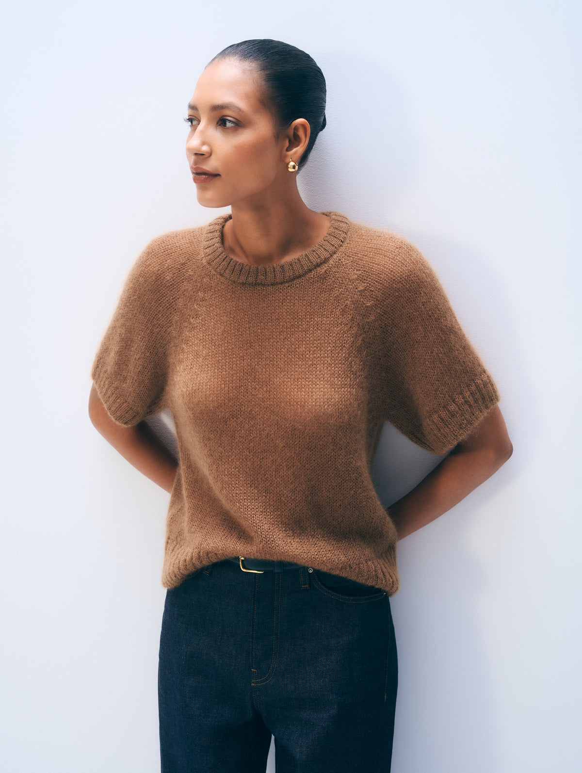A woman wearing a short-sleeve knit crewneck sweater in a warm camel brown, paired with dark blue jeans, standing against a light gray wall.