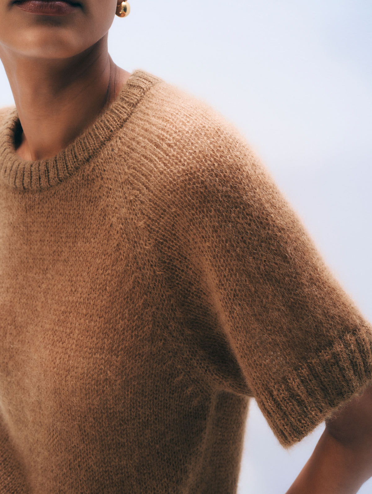 Close-up of a person wearing a short-sleeve knit sweater in a warm camel brown color, with a light blue sky background.