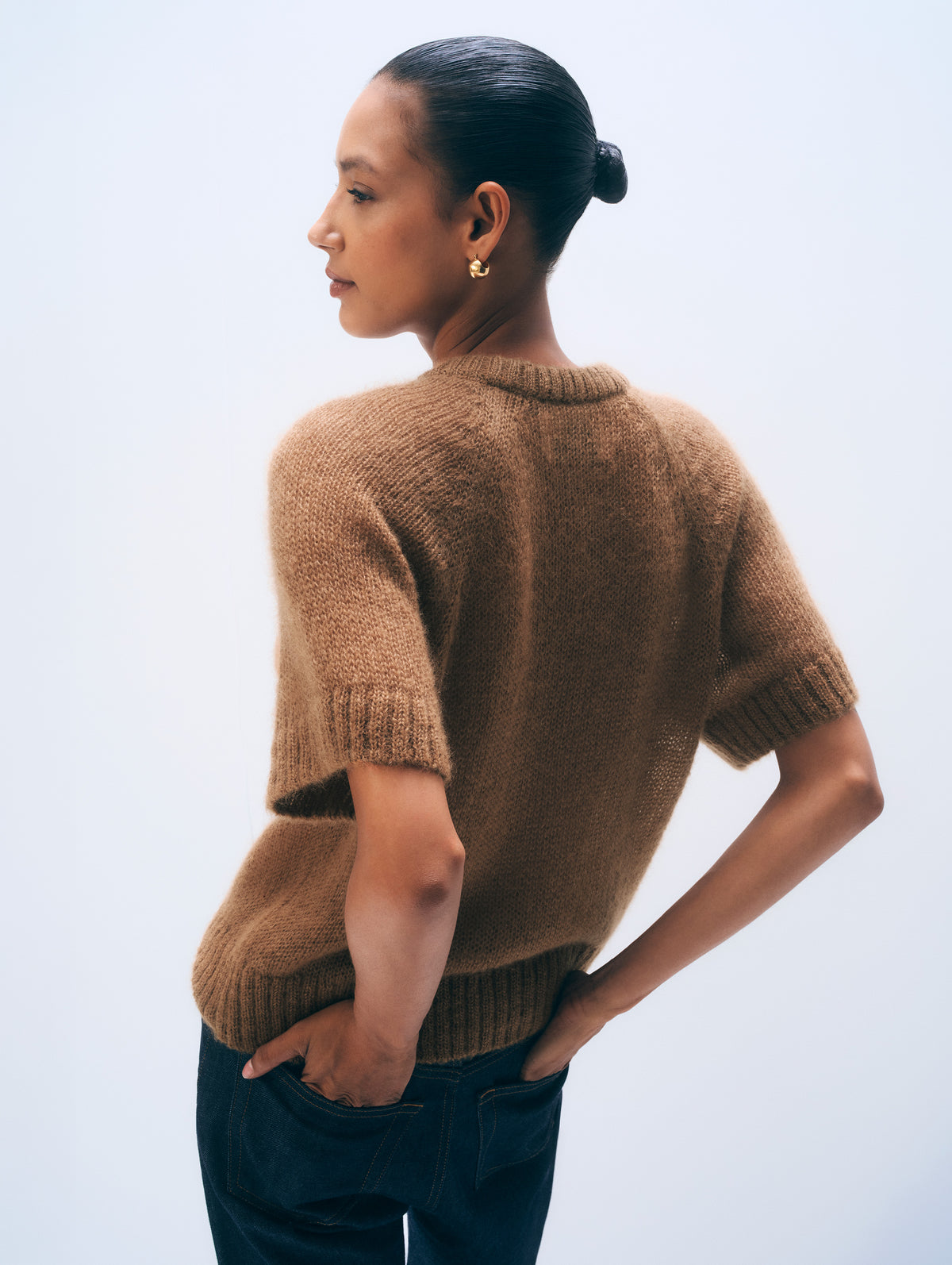 A woman shown from the back wearing a short-sleeve brown knit sweater and dark jeans against a light gray studio background.