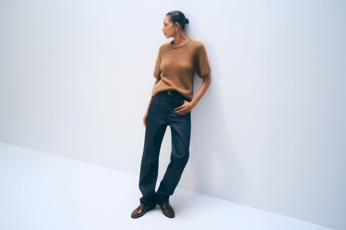 A model stands against a clean white corner wall wearing a short-sleeve brown knit sweater, dark navy trousers, and brown leather shoes.