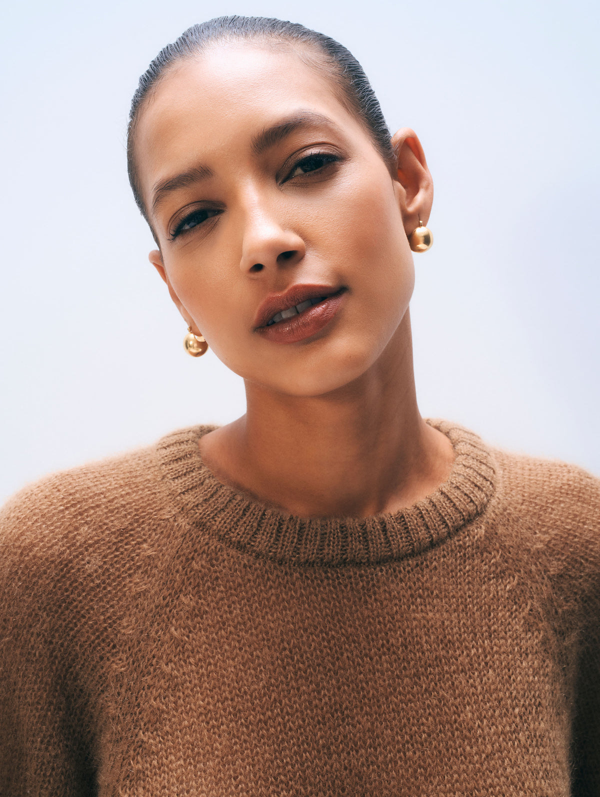 A close-up portrait of a woman with her hair pulled back, wearing a brown knit crewneck sweater and small gold hoop earrings against a light gray background.