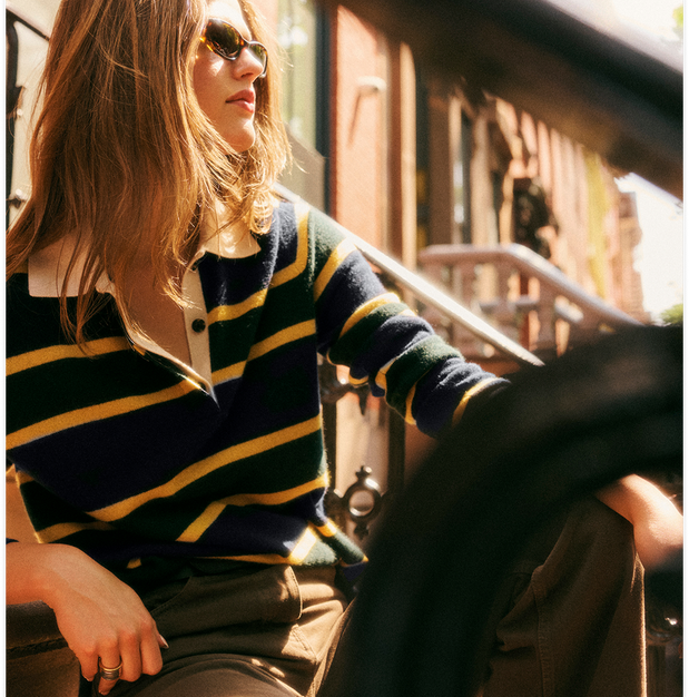 A woman wearing a navy and yellow striped knit polo sweater and brown pants, seated in a sunlit urban setting with a blurred dark foreground element in the foreground.