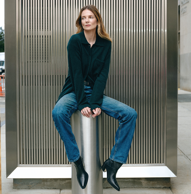 A woman sits on a cylindrical metal pedestal in front of a large slatted metal wall, wearing a dark green long-sleeve button-front jacket, blue jeans, and black ankle boots.