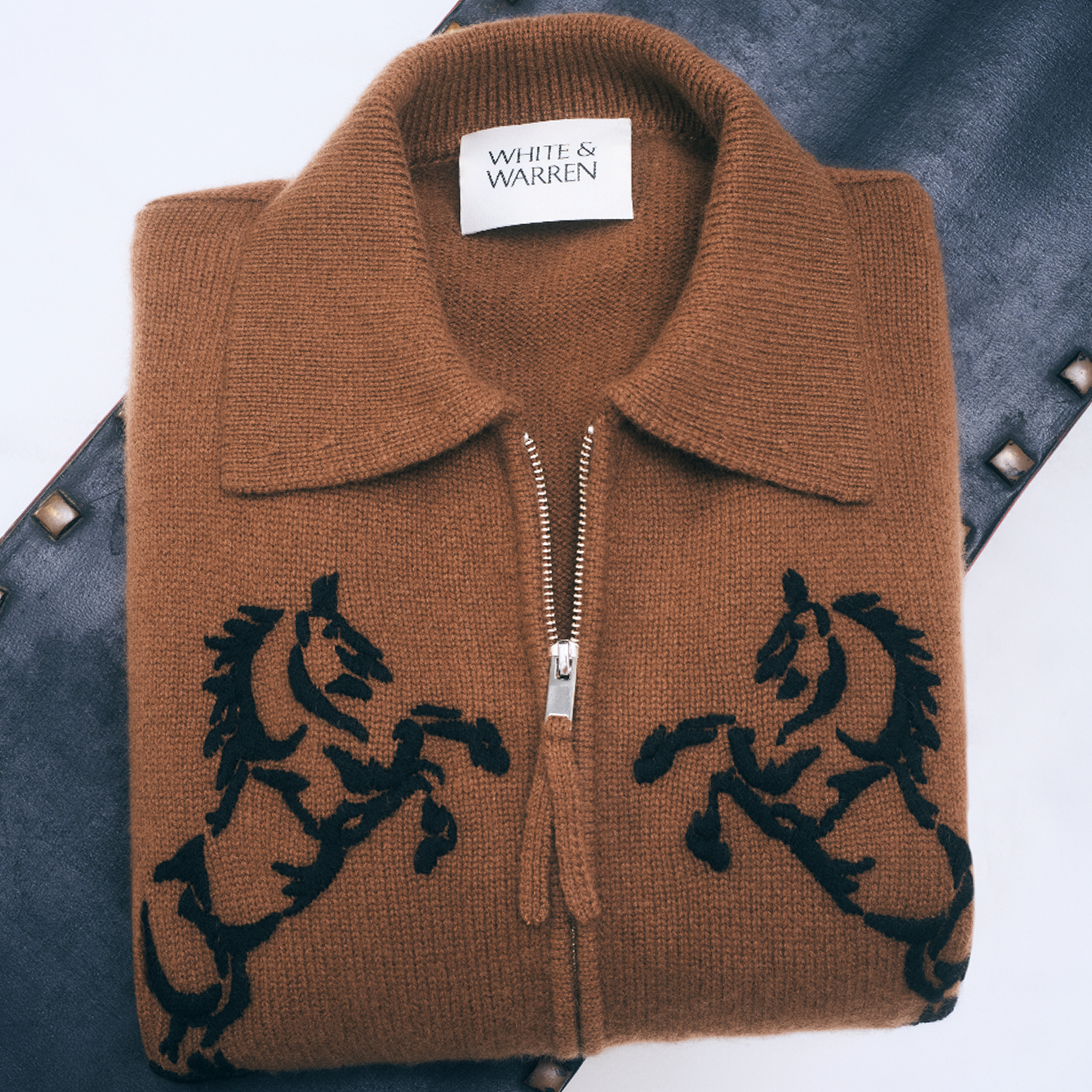 A folded brown knit zip-up sweater with black embroidered horse motifs on the front, displayed on a dark textured surface. The neck label reads 'WHITE & WARREN'.